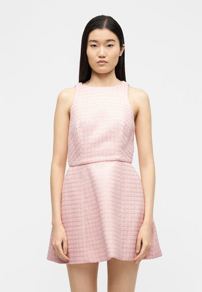 Νεαρή γυναίκα που φοράει ένα ροζ αμάνικο φόρεμα με υφή και φούστα σε σχήμα καμπάνας, στέκεται μπροστά σε ένα απλό λευκό φόντο.