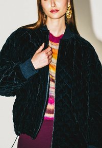 Bomber nero trapuntato con polsini a costine, cerniera frontale e texture morbida, indossato sopra un maglione multicolore.