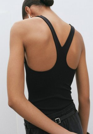 Personne aux cheveux courts portant un débardeur noir à dos nageur et un pantalon noir avec une ceinture, debout les mains derrière le dos.