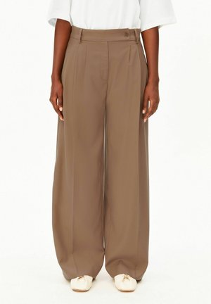 Personne portant un pantalon marron taille haute à jambes larges, un haut blanc et des chaussures plates écrues, se tenant droite devant un fond uni.