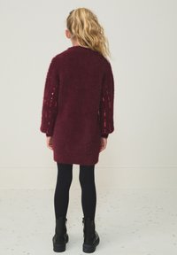 Burgundska pletena obleka z zamegljeno teksturo, dolgimi rokavi z bleščicami, kombinirana s črnimi nogavicami in gležnjarji.