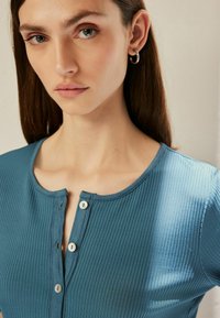 Teal geribbelde top met een ronde halslijn, voorzien van een knoopsluiting en korte mouwen. Gladde textuur, aansluitend ontwerp en subtiele glans.