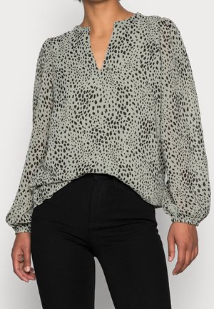 Femme portant un chemisier gris clair à taches noires irrégulières et manches longues bouffantes, associé à un pantalon noir taille haute.