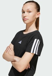 T-shirt noir à manches courtes en coton, avec des accents blancs à trois bandes sur les manches et un petit logo sur la poitrine.