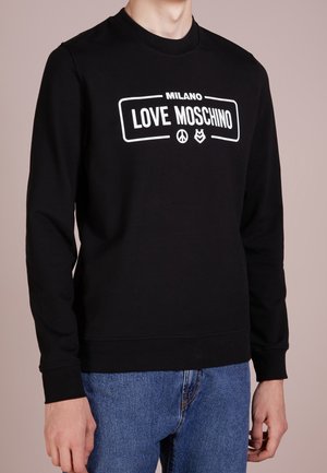 Osoba nosící černou mikinu s dlouhým rukávem s nápisem "MILANO LOVE MOSCHINO" a symboly, v kombinaci s modrými džínami.