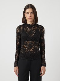 Top lungo in pizzo nero a maniche lunghe con motivi floreali, scollatura a onde e silhouette aderente, indossato con pantaloni neri.