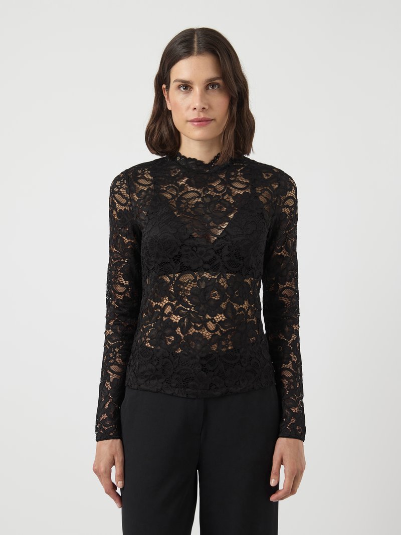 Top lungo in pizzo nero a maniche lunghe con motivi floreali, scollatura a onde e silhouette aderente, indossato con pantaloni neri.