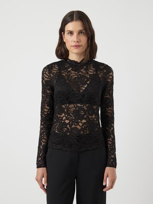 Top lungo in pizzo nero a maniche lunghe con motivi floreali, scollatura a onde e silhouette aderente, indossato con pantaloni neri.