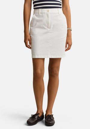 Frau trägt einen weißen knielangen Rock, schwarze Boots und ein marineblau-weiß gestreiftes Oberteil, steht mit entspannten Armen.