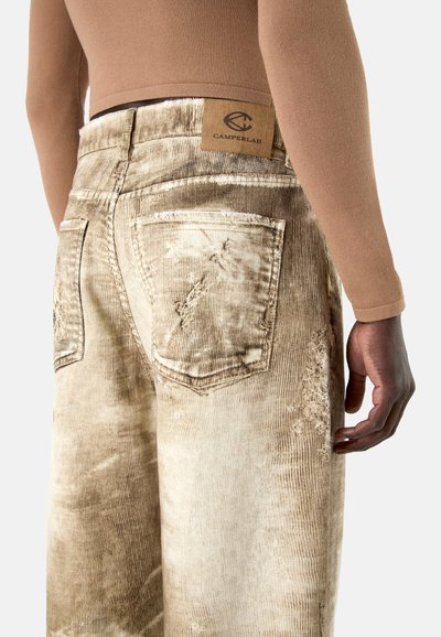 Vue arrière d'une personne portant un jean délavé beige avec des détails usés et un patch en cuir CAMPERLAB sur la ceinture.