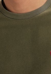 T-shirt à col rond en coton vert olive avec encolure côtelée, présentant un petit logo rouge sur la partie gauche de la poitrine. Texture douce et coupe décontractée.