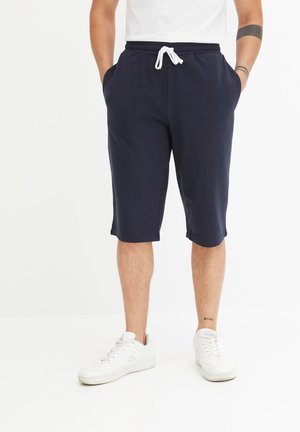 Pantaloni corti di cotone blu navy con vita elastica, cordoncino bianco, tasche laterali e taglio alla ginocchia. Indossati con scarpe da ginnastica bianche.