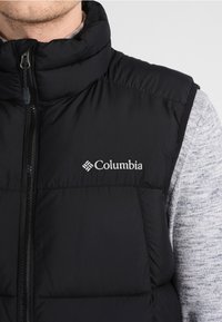 Svart pufferväst med hög krage, framzipper och Columbia-logotyp. Materialet är slätt med kviltade sektioner för isolering.