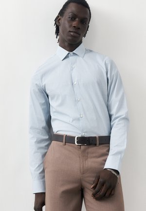 Άνδρας με σκούρο δέρμα που φοράει ανοιχτό μπλε καρό πουκάμισο με κουμπιά, μπεζ παντελόνι, μαύρη ζώνη και ασημένιο βραχιόλι, σε λευκό φόντο.