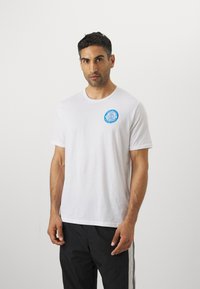 Balta kokvilnas T-kreklu ar apaļu izgriezumu un īsām piedurknēm, uz kura kreisajā krūtī ir izdrukāts zils apļveida logotips.