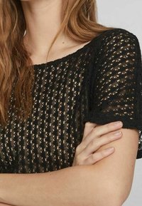 Top de crochet negro con un patrón texturizado, mangas cortas y un corte holgado. La tela tiene un diseño de tejido abierto con un borde en forma de concha.