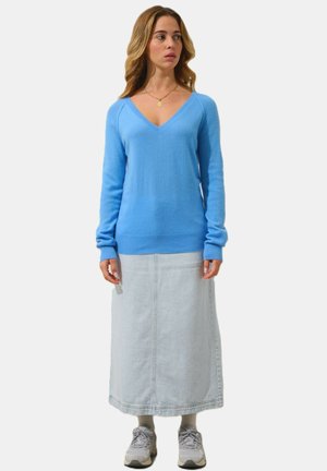 Hellblaue V-Ausschnitt-Sweater mit langen Ärmeln; kombiniert mit einem langen Denim-Maxikleid. Die Schuhe sind graue Sneaker. Einfaches Design und strukturierter Stoff.