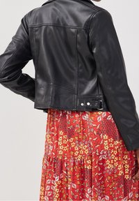 Veste cropped en cuir noir avec une texture lisse, grands revers et accents à bouton-pression, portée sur une robe rouge à imprimé floral.