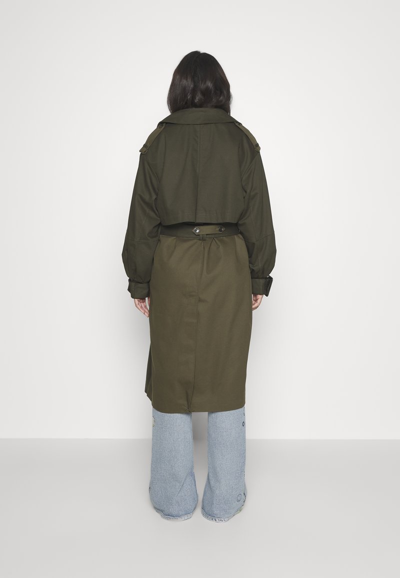 Olivgrüner Trenchcoat mit tailliertem Gürtel, weiten Ärmeln und Schulterklappen, kombiniert mit hellblauen, weit geschnittenen Jeans.