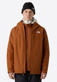 Chaqueta marrón resistente al agua con cremallera frontal, capucha y forro de polar, superpuesta a una chaqueta interior ligera. Se incluye un gorro negro.
