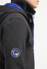 Svart jacka med hög krage och blå detaljer, samt en rund logotyp-patch på vänster ärm. Slät textur, midje-lång design.