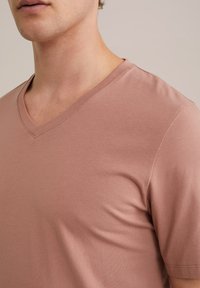 V-alakú nyakkivágású, rövid ujjú póló halvány rózsaszínben, puha pamutból készült. Simán textúrája és tiszta varrásai vannak a nyakkivágás mentén.