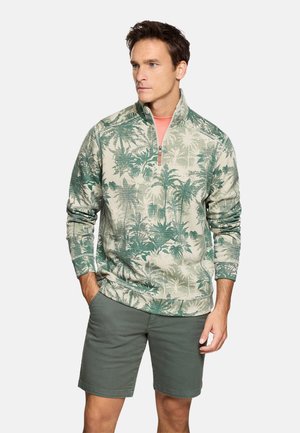 Hombre vistiendo un pulóver verde con estampado tropical y cremallera parcial, junto con pantalones cortos verde oscuro, de pie con una mano en el bolsillo, mirando hacia un lado.