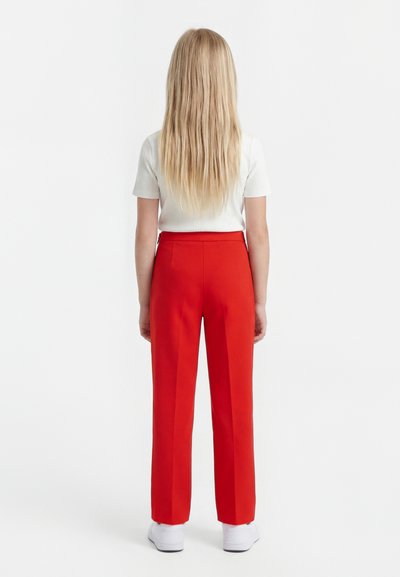 Enfant aux longs cheveux blonds, dos à l’objectif, portant un haut blanc à manches courtes, un pantalon rouge vif et des baskets blanches sur fond blanc.