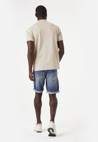Licht beige polo shirt met korte mouwen, gecombineerd met opgerolde blauwe denim shorts. Persoon in witte sportschoenen, van achteren bekeken.