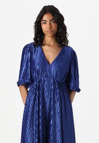 Blue, pleated satin dress with a deep V-neck, short puff sleeves, and gathered waist. Features ruffled accents at sleeve hems.