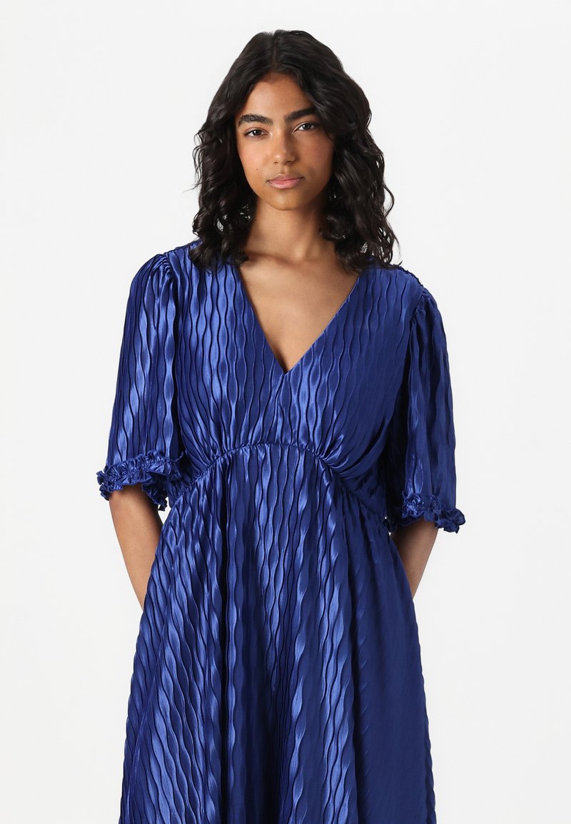 Niebieska, plisowana satynowa sukienka z głębokim dekoltem w serek, krótkimi bufkami oraz marszczoną talią. Posiada falbany przy brzegach rękawów.