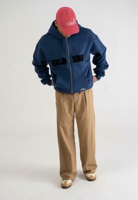 Felpa con zip navy con "BERLIN SOH" in testo nero, abbinata a pantaloni larghi beige e sneakers beige. Un cappellino rosso completa l'outfit.