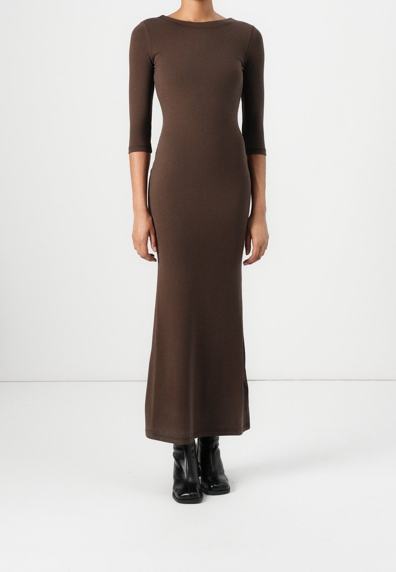 Femme portant une longue robe marron près du corps avec des manches trois-quarts et des bottines noires, debout devant un fond blanc uni.