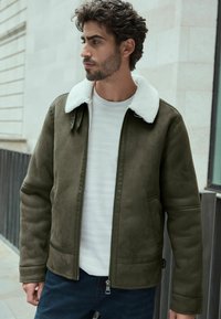 Chaqueta verde olivo con un cuello de piel blanca, con cremallera en la parte delantera y dos bolsillos laterales. Llevada sobre una camiseta blanca de canalé y pantalones de mezclilla oscuros.