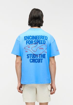 Osoba nosiaca modré tričko s nápisom „Navrhnuté pre rýchlosť“ a mapou pretekárskej dráhy označenou ako „Rakúska Grand Prix“ na chrbte.