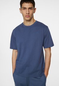 Sininen puuvillapaita, jossa on lyhyet hihat, pyöreä kaula-aukko ja pieni logo vasemmassa rinnassa. Sileä pinta, rento istuvuus.