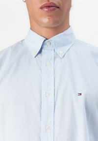 Mann iført lys blå mønstrete skjorte med knapp og liten brodert logo på brystet, med knappet krage, mot hvit bakgrunn.