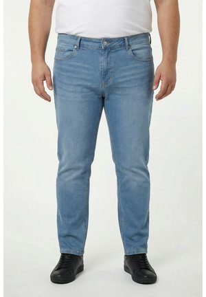 Mann trägt hellblaue Jeans, schwarze Sneakers und ein weißes Hemd, steht vor einem einfarbigen hellen Hintergrund.