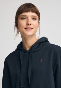 Huvtröja i mörk marinblått tyg, med en framficka i känguru-stil och en liten röd broderad logotyp på bröstet.