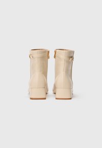 Bottines beiges en cuir lisse, avec un talon bloc et des fermetures éclair latérales ornées de détails en or. Semelles plates texturées.