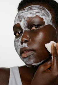 Ansiktsrengöringsprocess med vit kräm applicerad på mörk hud. En person håller en liten vit duk mot sin kind och bär en grå linne.