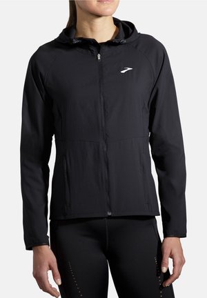 Femme portant une veste noire à capuche avec fermeture éclair et des leggings de sport noirs, debout, bras détendus le long du corps.