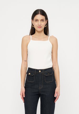 Γυναίκα με μακριά σκουρόχρωμα μαλλιά, που φοράει λευκό αμάνικο τοπ και ψηλόμεσα σκουρό μπλε τζιν με μπροστινές τσέπες με πάτωμα, στέκεται μπροστά σε λευκό φόντο.