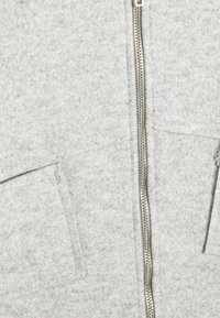 Sudadera con cremallera gris hecha de una tela suave y texturizada; cuenta con dos bolsillos frontales y una cremallera plateada que recorre el centro.