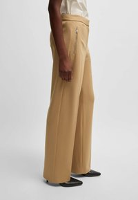 Beige wide-leg trousers with a smooth texture, featuring a side zipper pocket and a high waist, paired with pointed black heels.