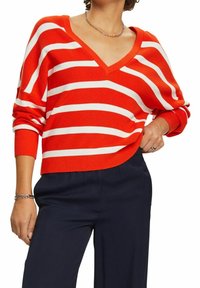 Rot-weiß gestreifter, gestrickter Pullover mit V-Ausschnitt, überschnittenen Schultern und gerippten Bündchen, kombiniert mit dunkelblauen Hosen.