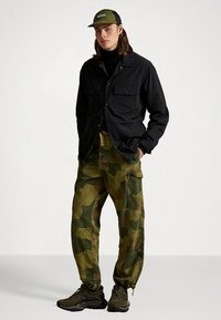 Czarna koszula na guziki, oliwkowe wojskowe spodnie typu cargo, czarne trampki i zielona czapka. Charakterystyczne detale to kieszenie na piersi i luźny krój.