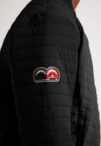 Giacca nera trapuntata con un prominente patch logo in rosso, nero e bianco sulla spalla sinistra. Tessuto testurizzato con chiusura a zip.