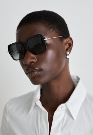 Black oversized sunglasses with a geometric frame, gold accents on the arms, worn with a white collared shirt, highlighting sleek design elements.