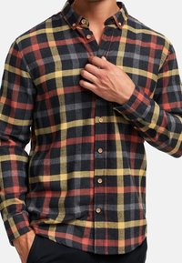 Homme portant une chemise à carreaux multicolore noire, rouge, jaune et grise, boutonnée, avec une main touchant la poitrine près du col.